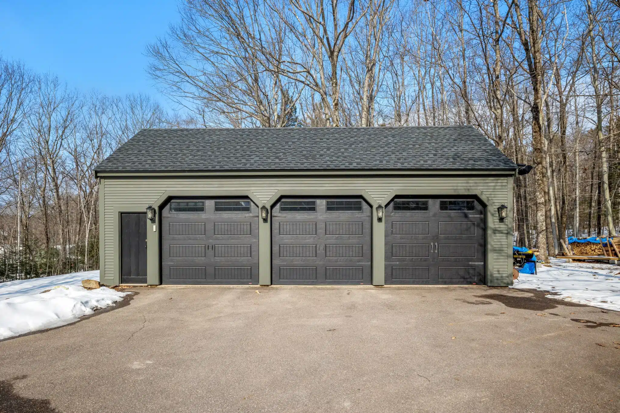 Garage triple isolé avec trois portes en bois foncé sur une allée goudronnée, entouré par une forêt en hiver.