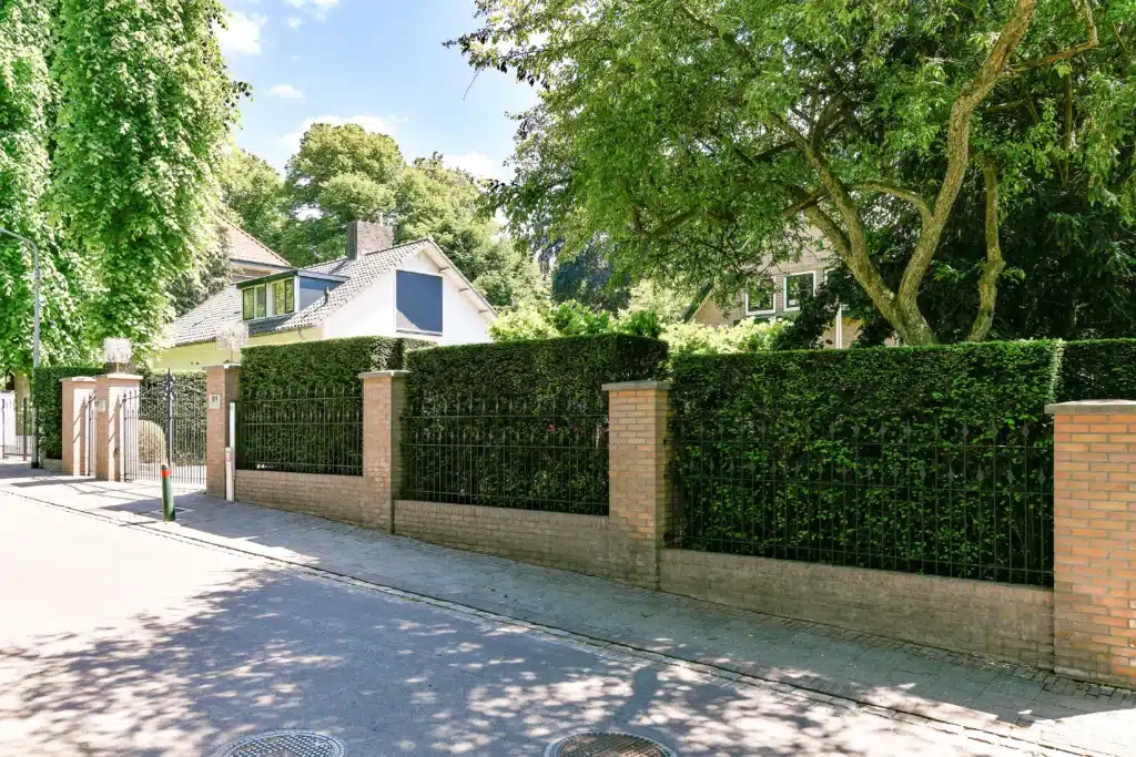 Muret de jardin avec entrée en briques et portail en métal, entouré d'une haie dense et d'un grand arbre.