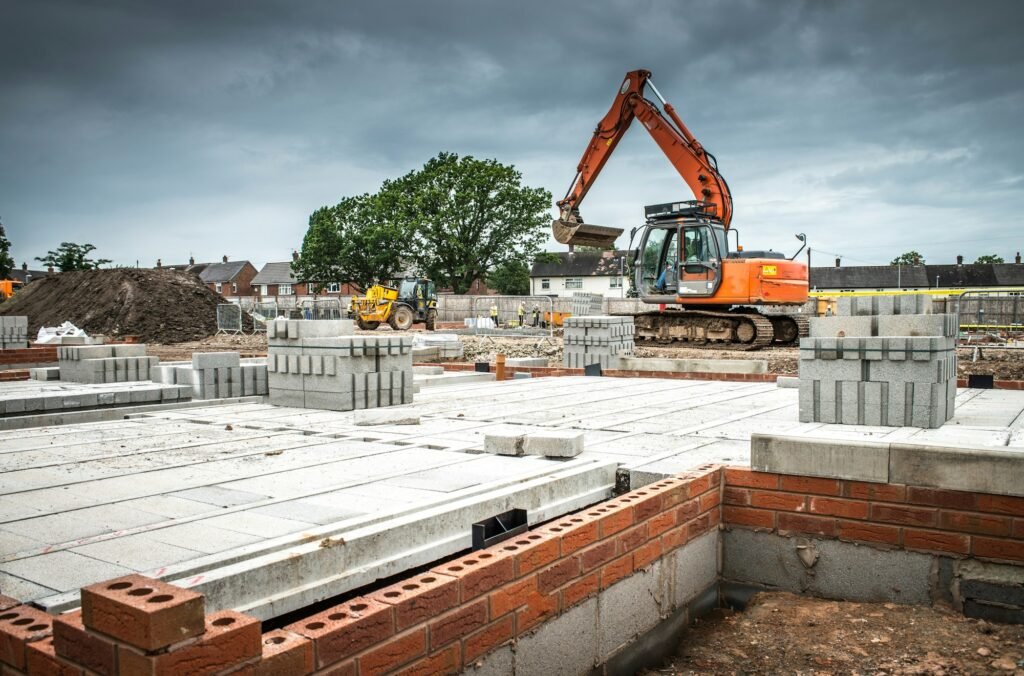 Un chantier de construction animé avec une grande excavatrice déplaçant de la terre à proximité de piles de blocs de béton et de renforts en acier.