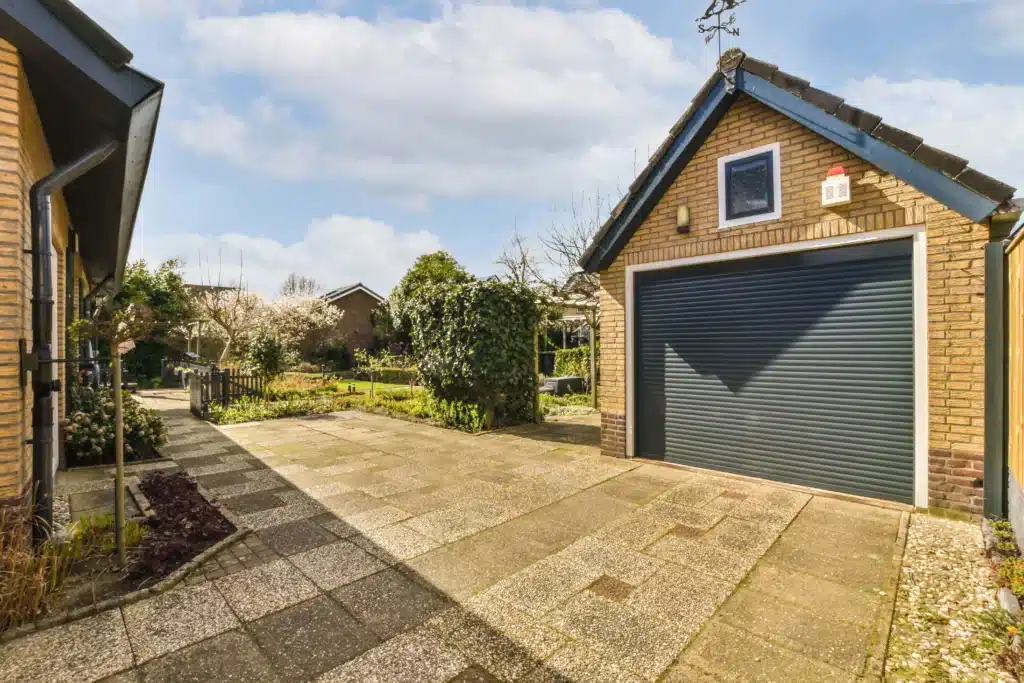 Garage détaché en brique avec porte en métal gris, situé à côté d'un jardin fleuri sous un ciel partiellement nuageux.