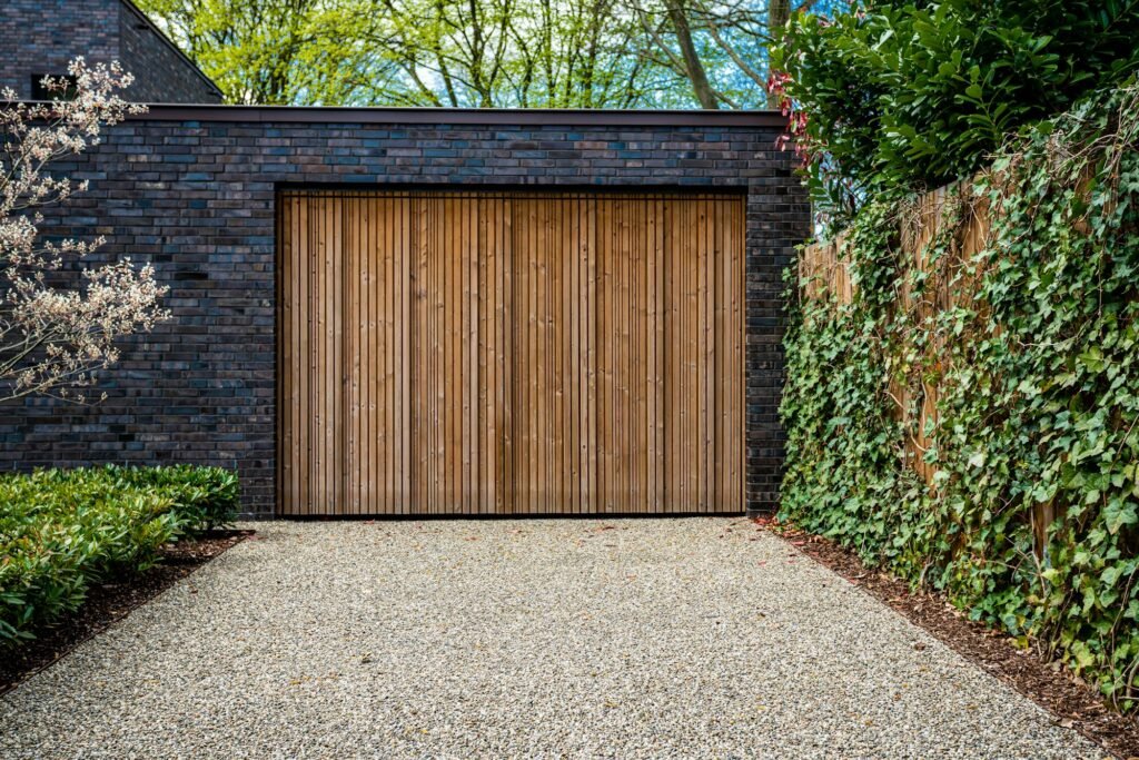 Garage résidentiel en brique noire avec une grande porte en bois, entouré de végétation et d'une allée en gravier.